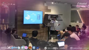 A speaker stands in front of an audience inside a modern event space during the AWS Student Builder Group Celestial Supernova event. The speaker, a young man in a white button-down shirt, is holding a microphone and gesturing with his left hand while presenting from a laptop. A large TV mounted on a wooden accent wall behind him displays a diagram of the client-server model, showing a laptop labeled "Client" sending a "Request" arrow to a rack of servers labeled "Server", which sends a "Response" arrow back. In the foreground, several attendees are sitting at tables, focused on the presentation with their own laptops open. The image features the event's standard digital watermarks and partner logos along the borders.